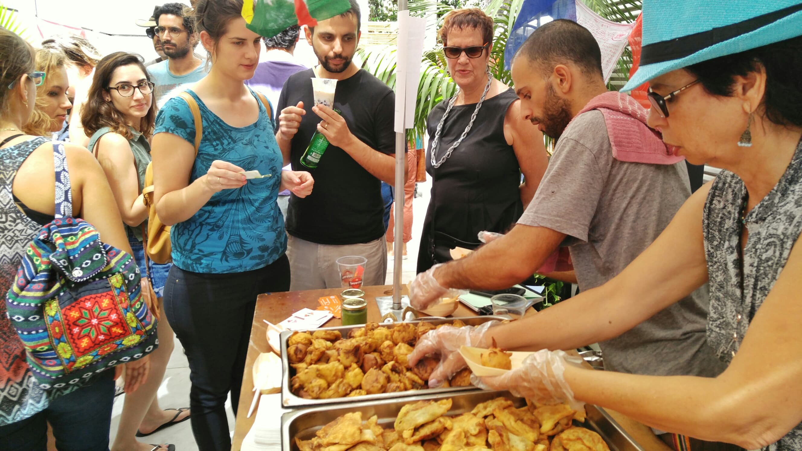 ידיים בכפפות מגישות סמוסות ופקורה מדוכן אוכל ביריד, ולפני הדוכן תור של לקוחות