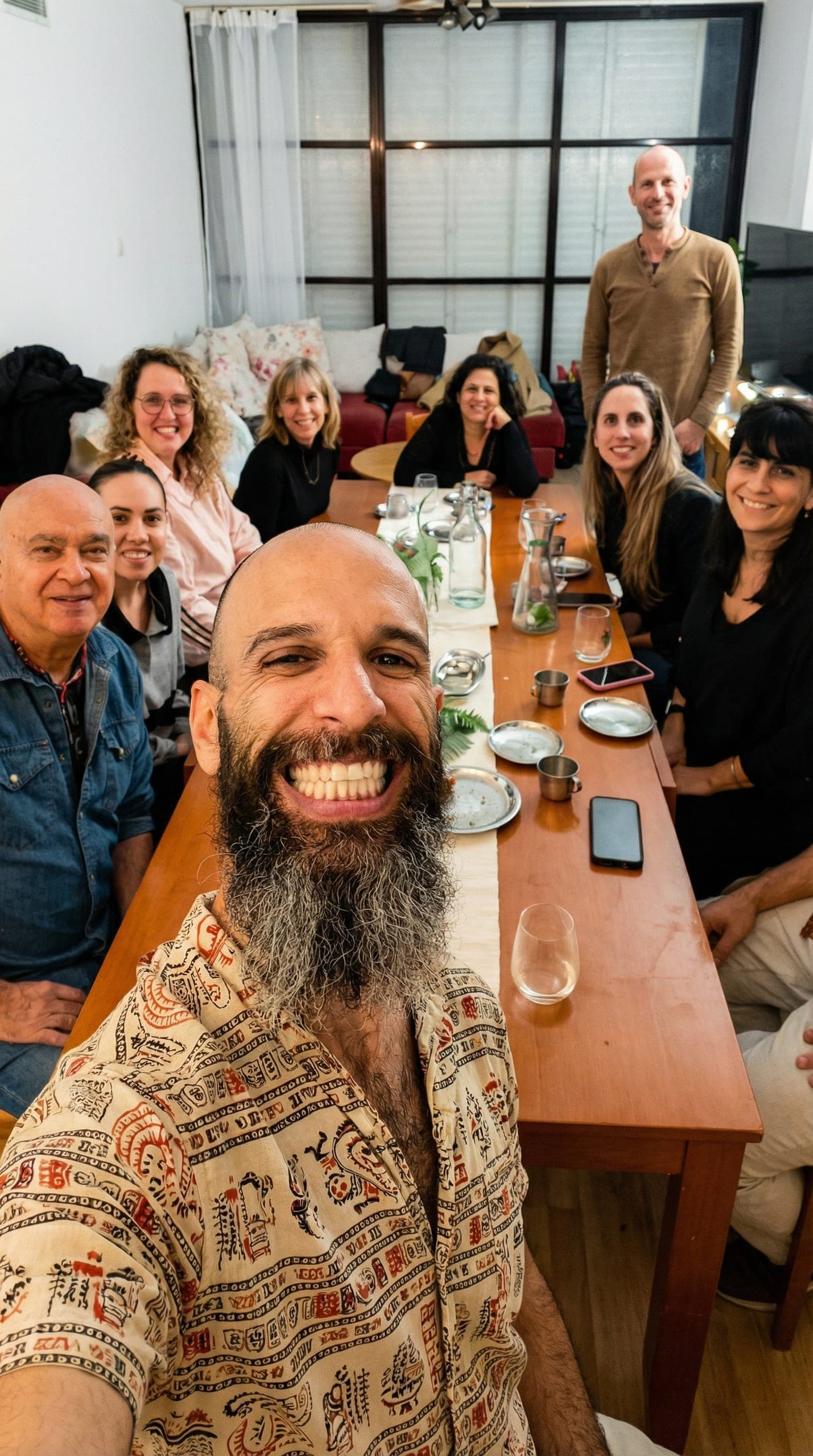 קבוצת אנשים מחייכים סביב שולחן אוכל מעץ, נהנים מארוחה משותפת באווירה חמה וביתית. במרכז התמונה גבר עם זקן ארוך מחייך למצלמה.
